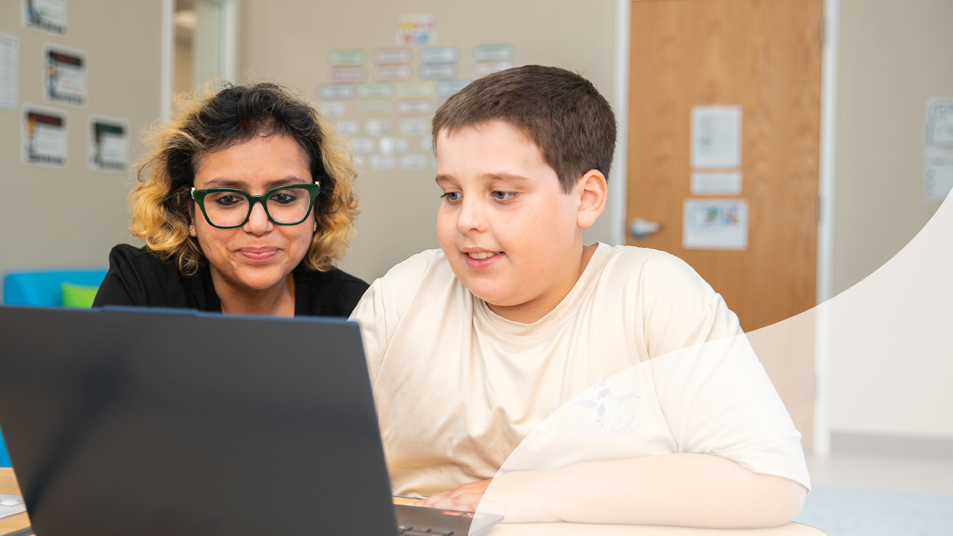A woman and a boy sit at a table looking at a laptop in a room with posters on the wall.