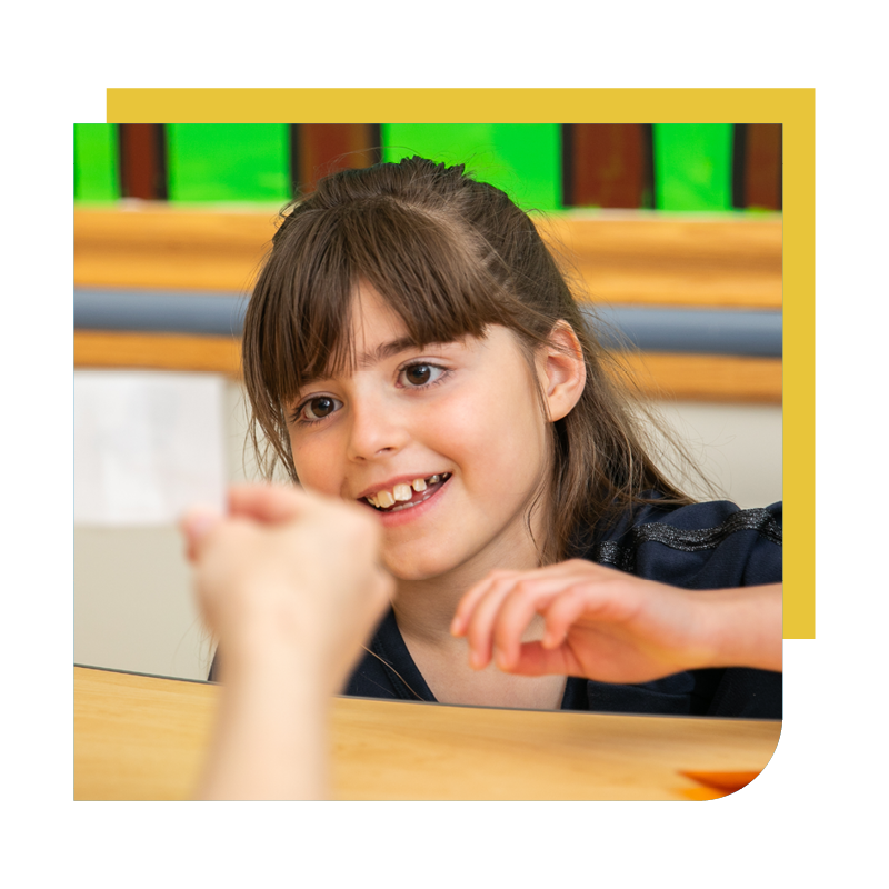 A young girl with brown hair and a gap-tooth smile sitting at a wooden table, engaged in a conversation or activity with an unseen person.