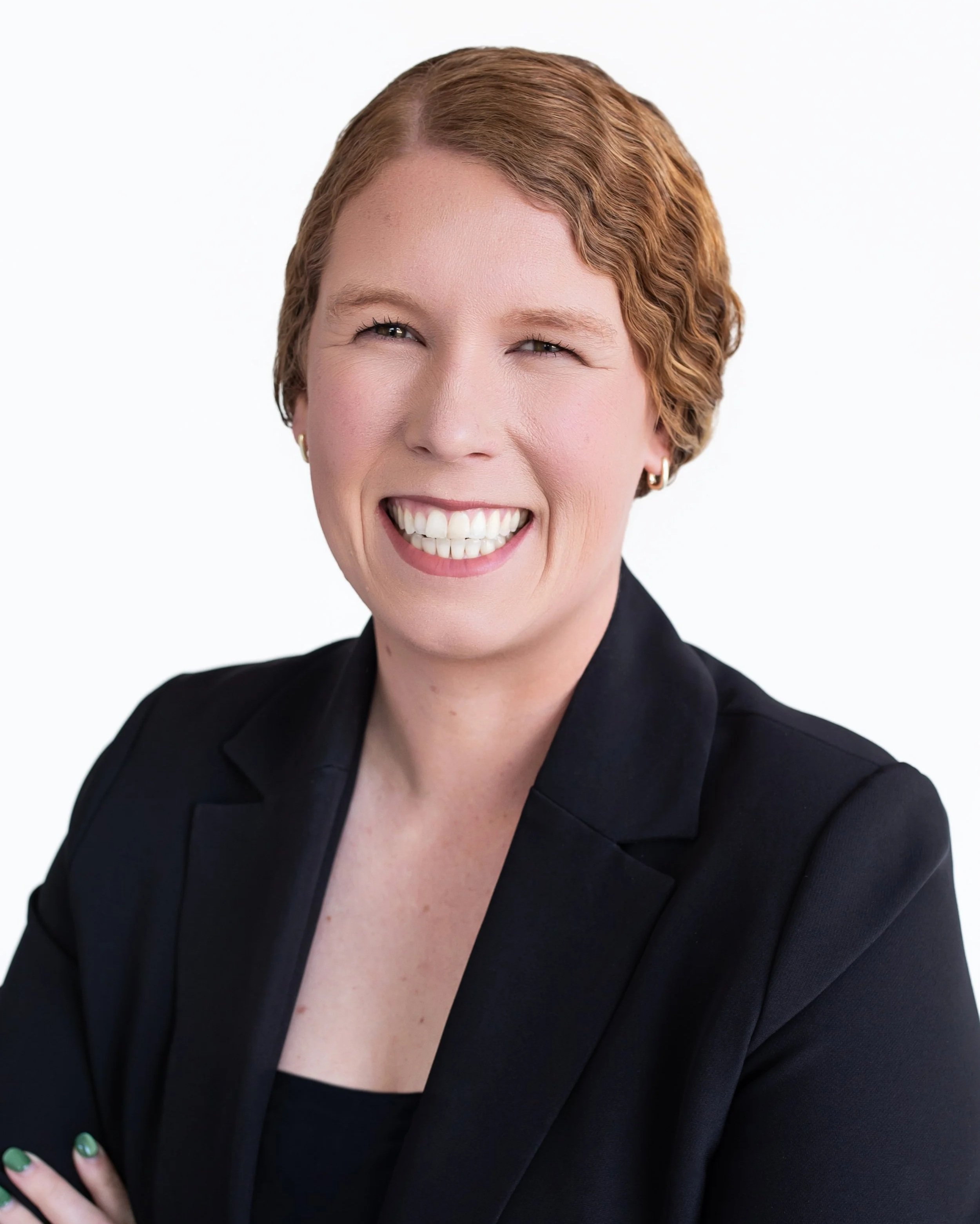 Close-up of a smiling woman with short red hair, wearing a black blazer, against a plain white background.