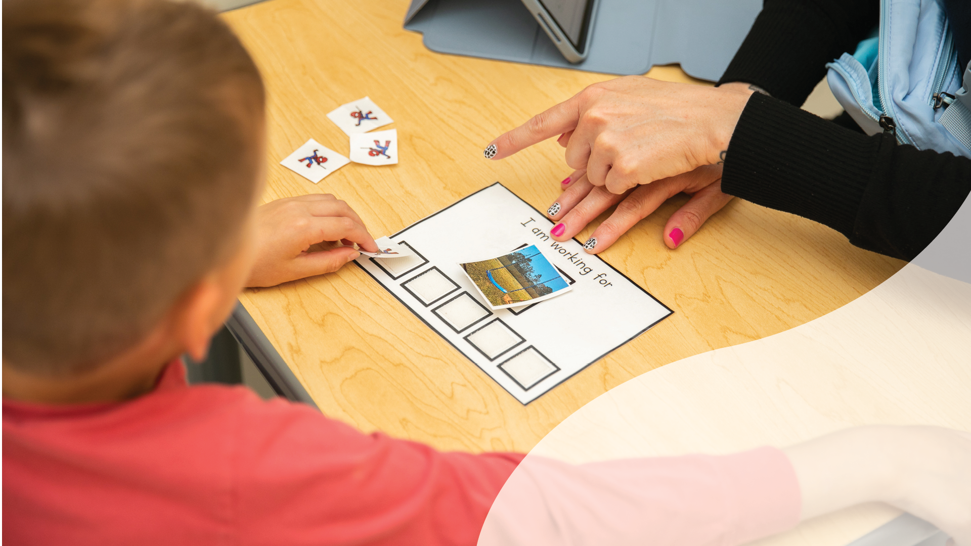 A teacher and a student working together on a visual activity at a classroom desk, with the teacher pointing to a picture of a park on a worksheet that says 'I am working for' and has empty boxes for photos or words.