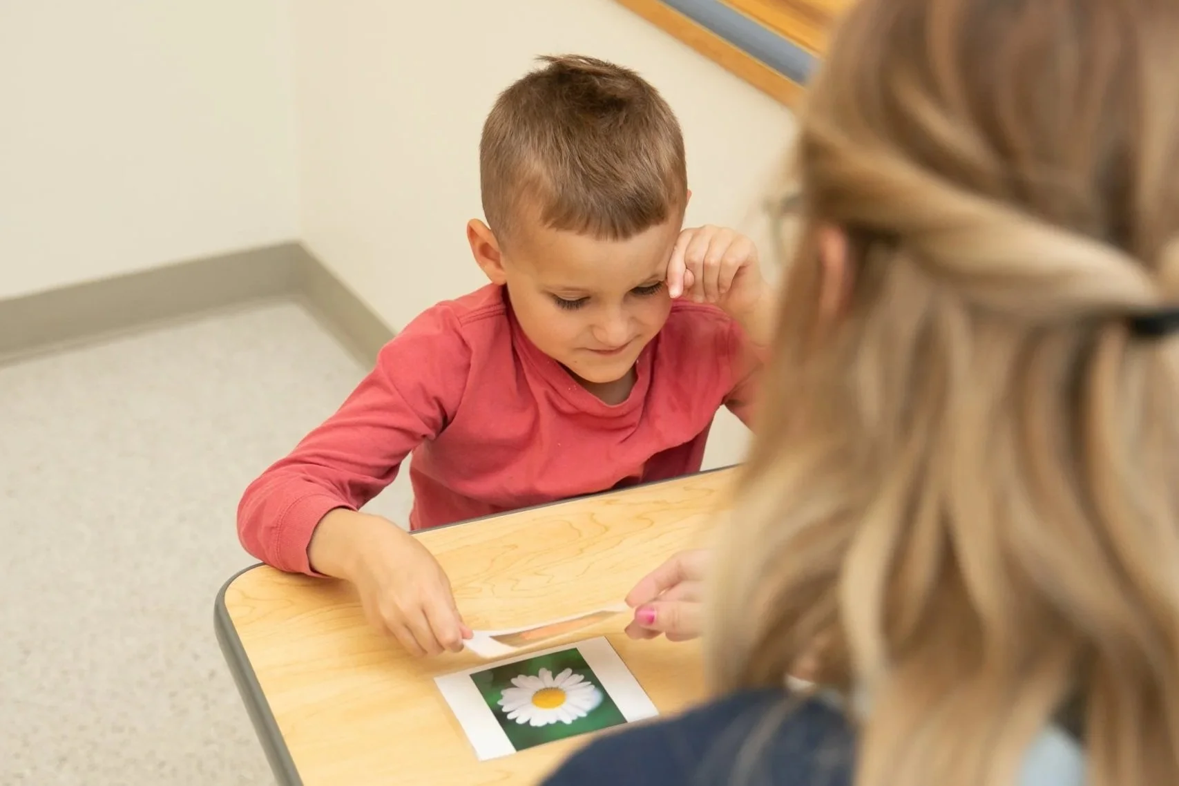 A young boy in a red shirt sitting at a table, concentrating as he points to a picture of a white daisy on a paper in front of him. An adult woman with long, blond, curly hair is sitting across from him, observing.
