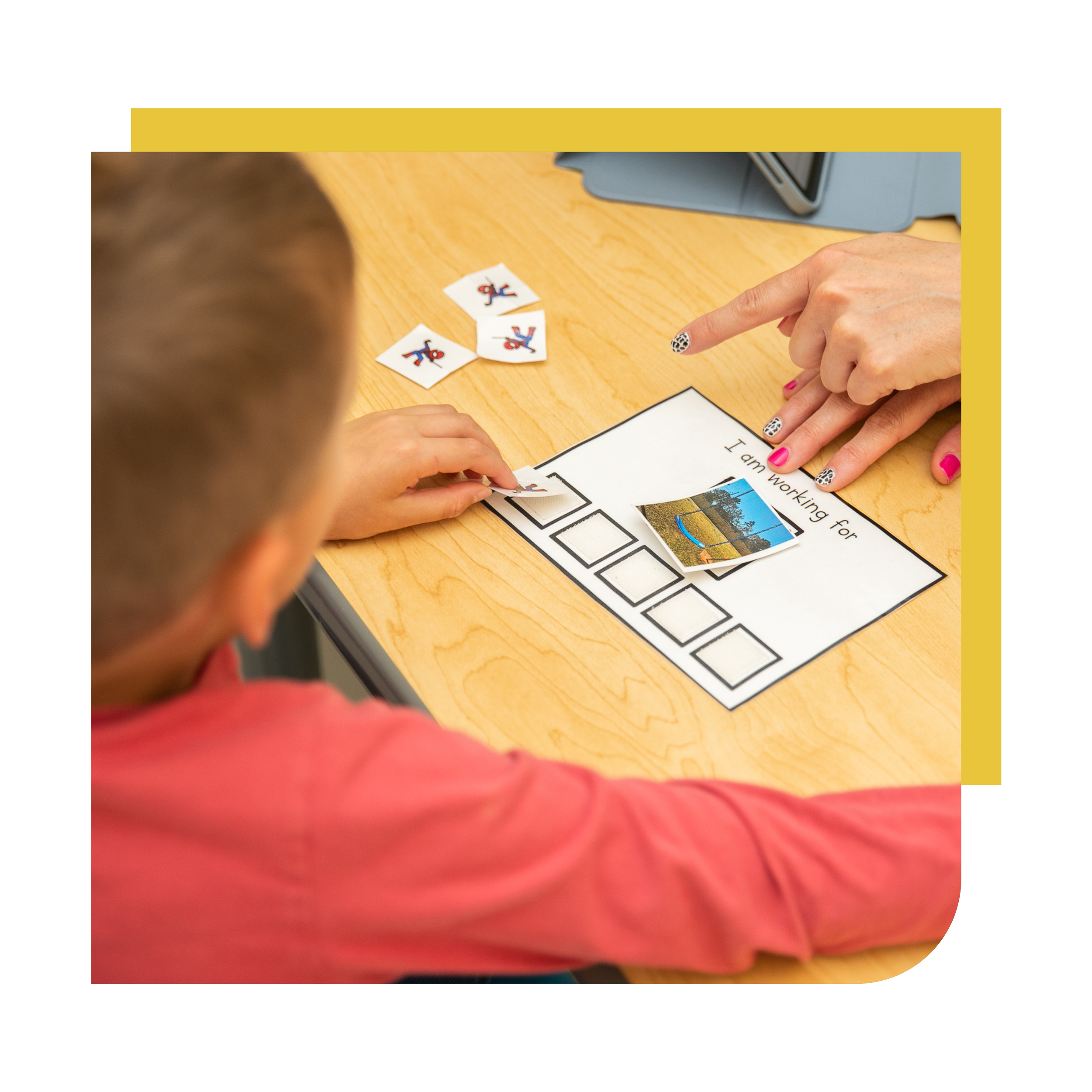 Close-up showing playing cards on a wooden table, with a woman's hand pointing at the cards, and a cardboard cutout of a blue and orange figure in the background.