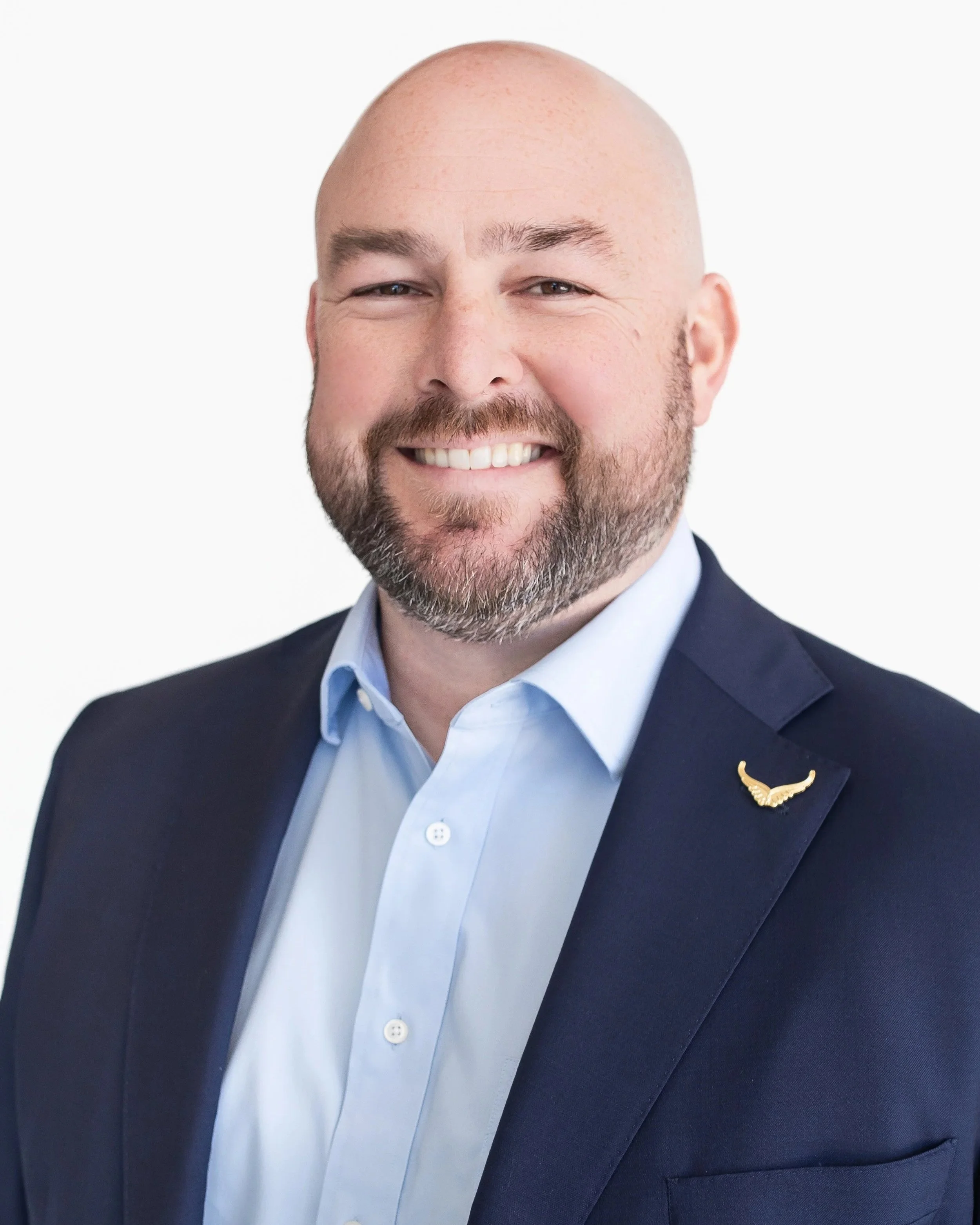 A smiling man with a beard, wearing a dark blazer and light blue dress shirt, with a gold wing lapel pin, against a plain white background.