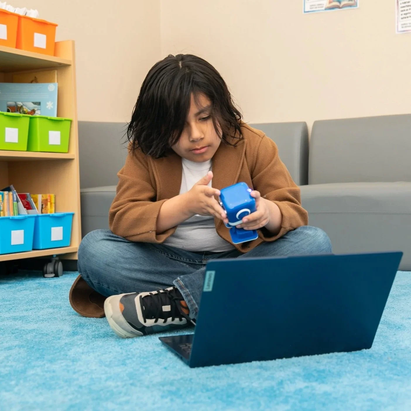 A young boy sitting cross-legged on a blue carpet in a classroom, holding a small blue toy cloud, with a black laptop in front of him, shelves with colorful bins and books in the background.