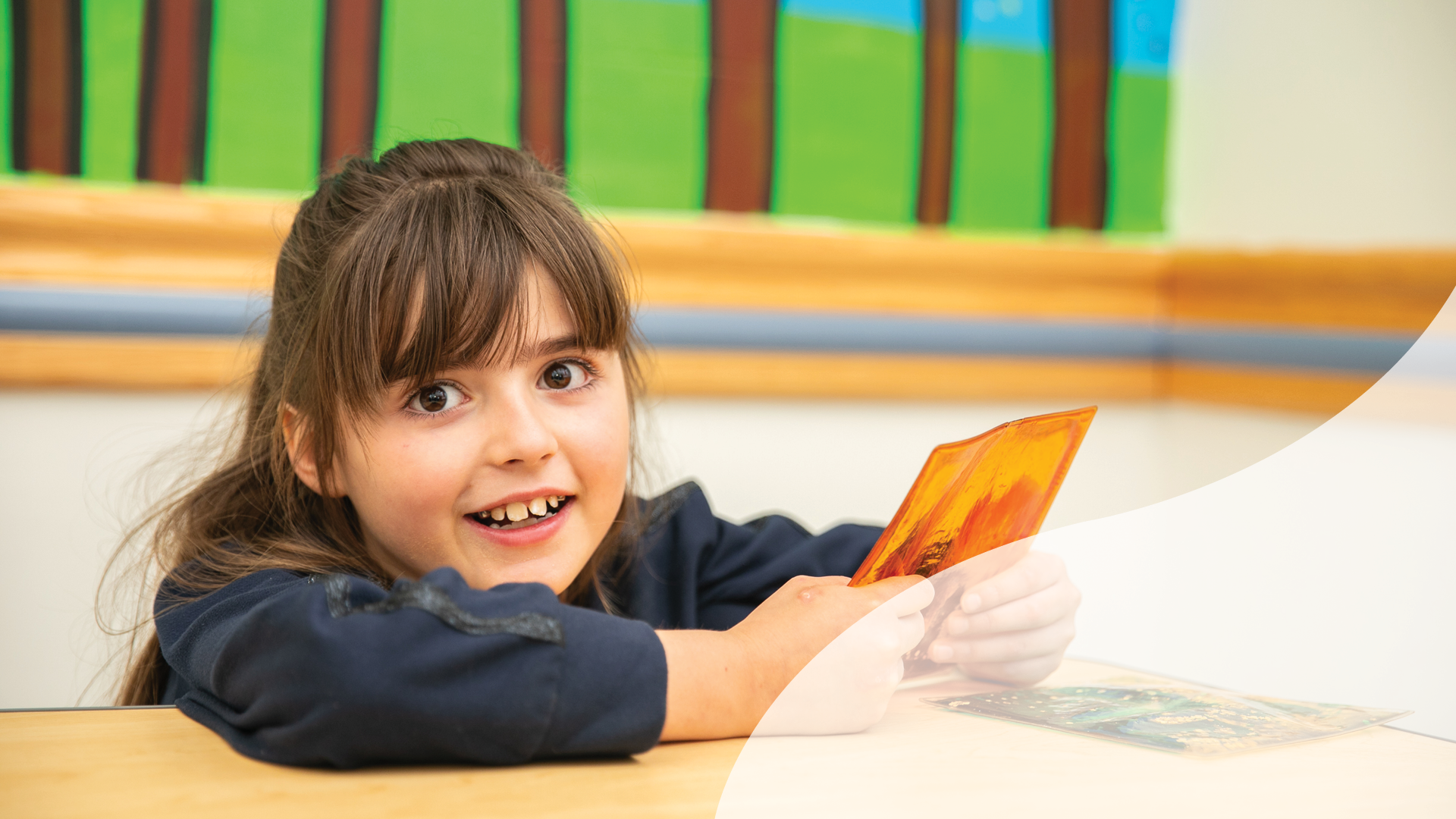 A young girl with long brown hair, smiling, holding an orange card, sitting at a school desk in a classroom with colorful decorations in the background.