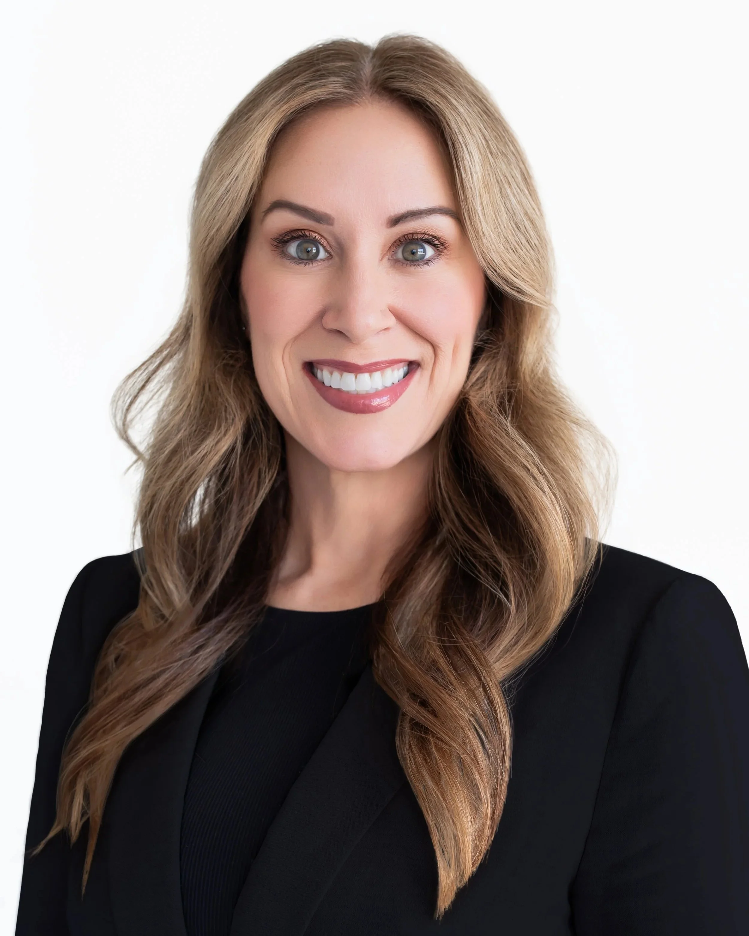 A woman with long, wavy, light brown hair smiling, wearing a black top, against a white background.