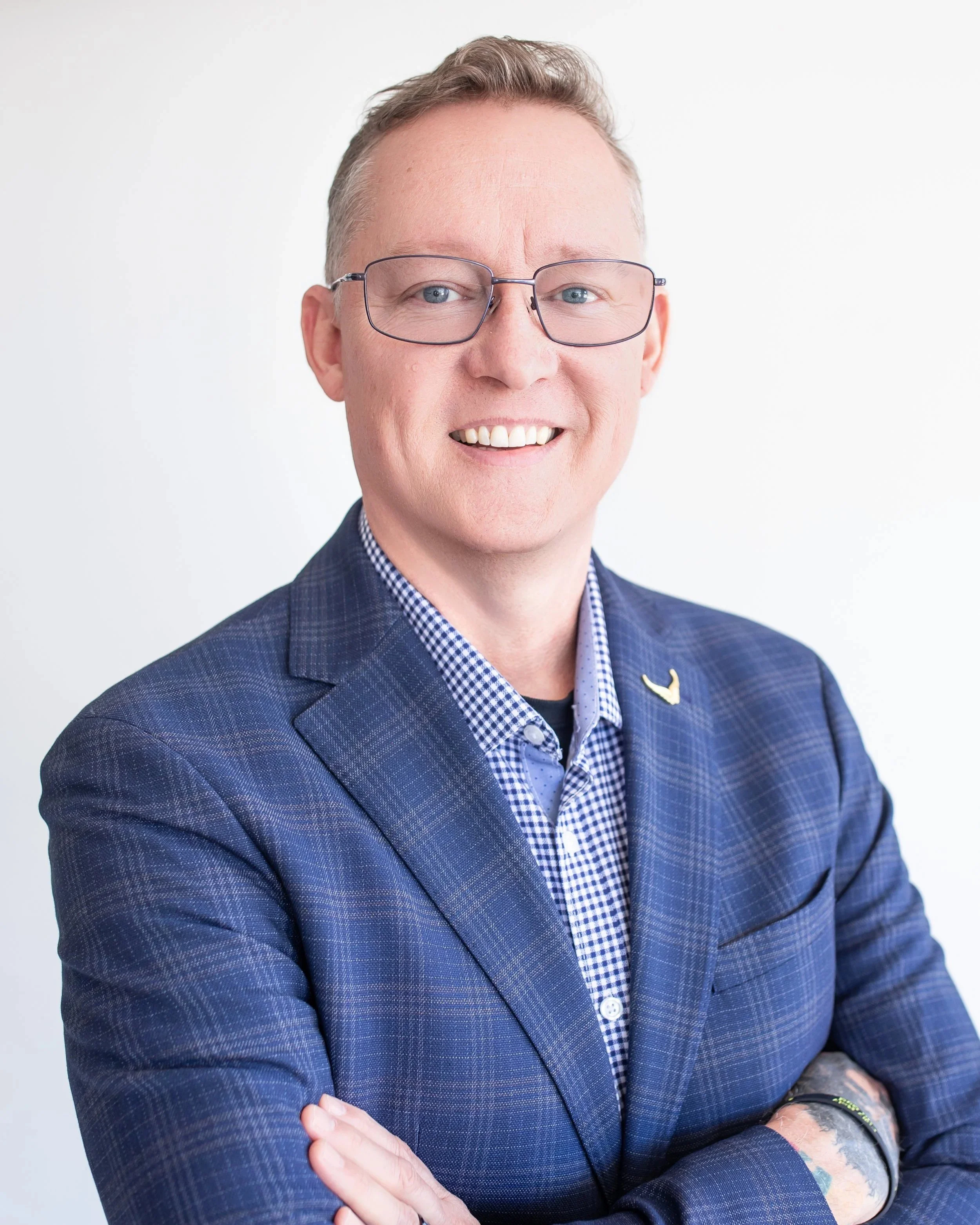 Close-up of a smiling man wearing glasses and a blue checked blazer with arms crossed, standing against a white background.