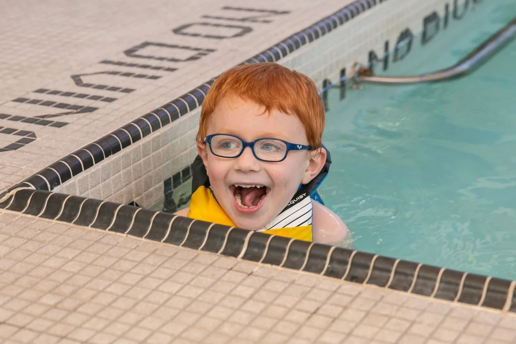A young boy with red hair, glasses, and a wide smile in a swimming pool, sitting near the edge with his mouth open in excitement.