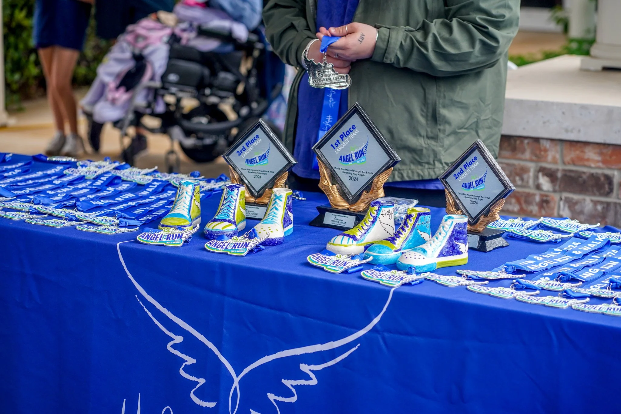 Table with blue tablecloth displaying medals, trophies, and decorated shoes for a race event, with people and a stroller in the background.