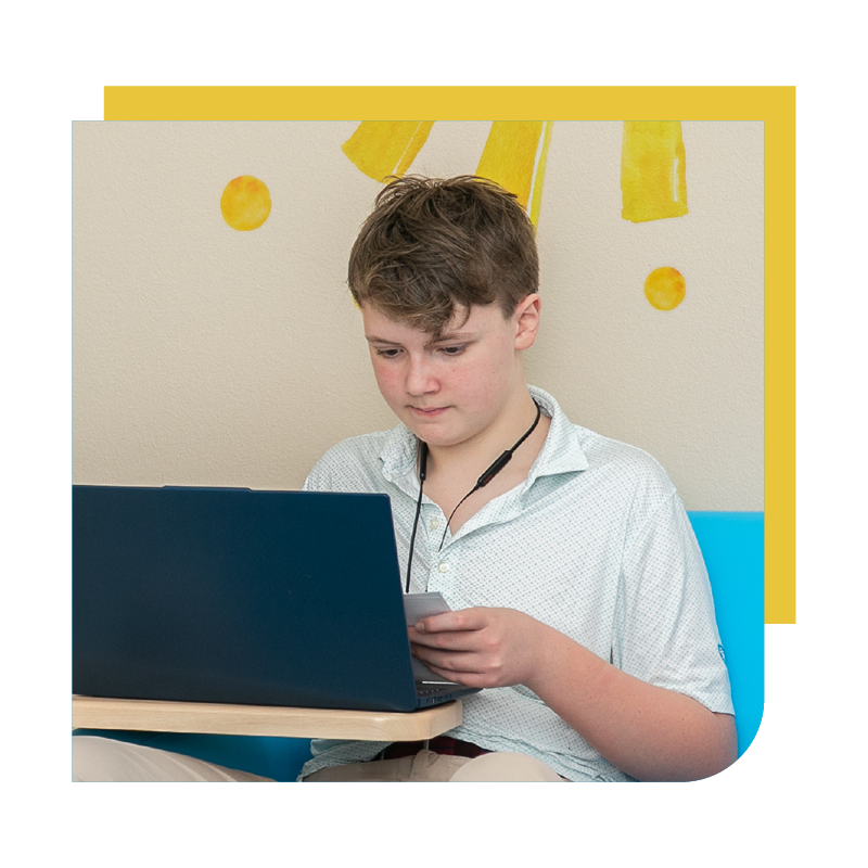 A young boy sitting at a table looking at his phone with a laptop open in front of him, wearing earphones, in a room with yellow and colorful wall decorations.