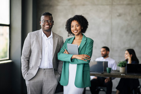 Two professionals, a man and a woman, standing and smiling in an office with other employees working in the background.