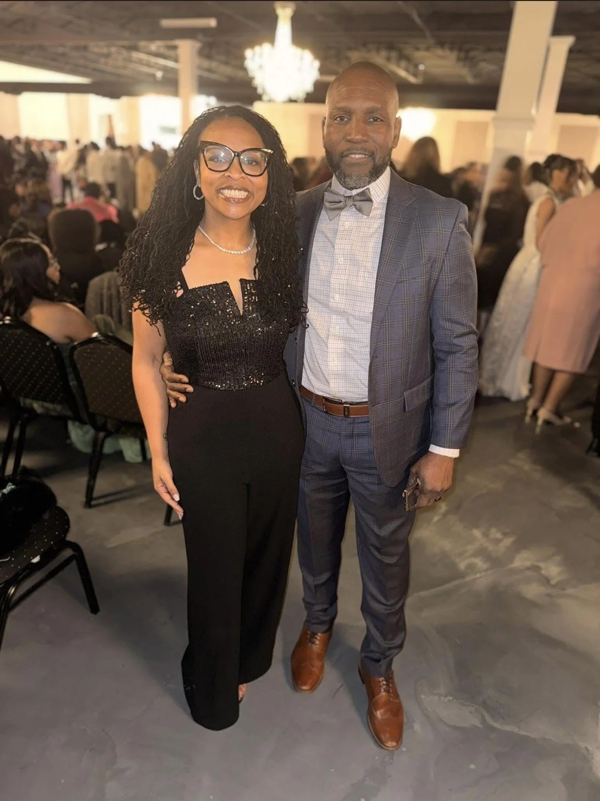 A man and woman dressed in formal attire posing together at an indoor event, with people and tables in the background.