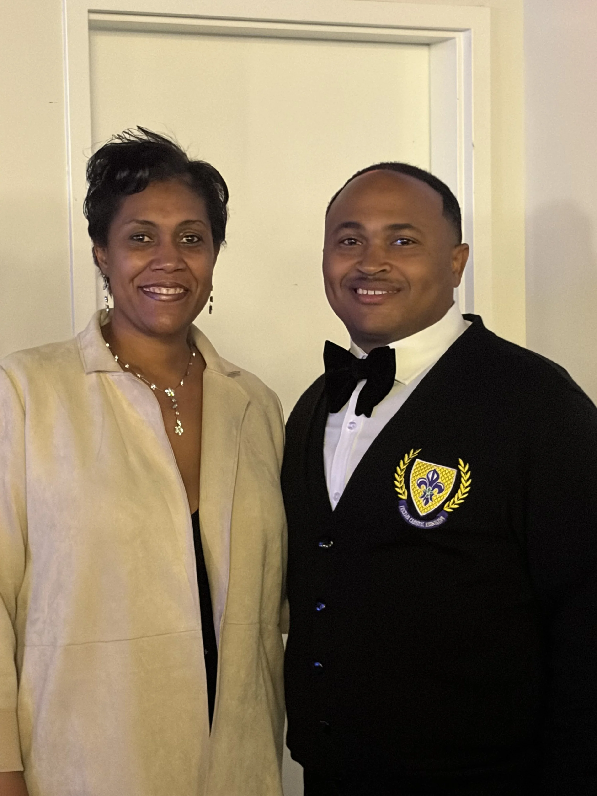 A woman and a man dressed formally, standing indoors, smiling at the camera.
