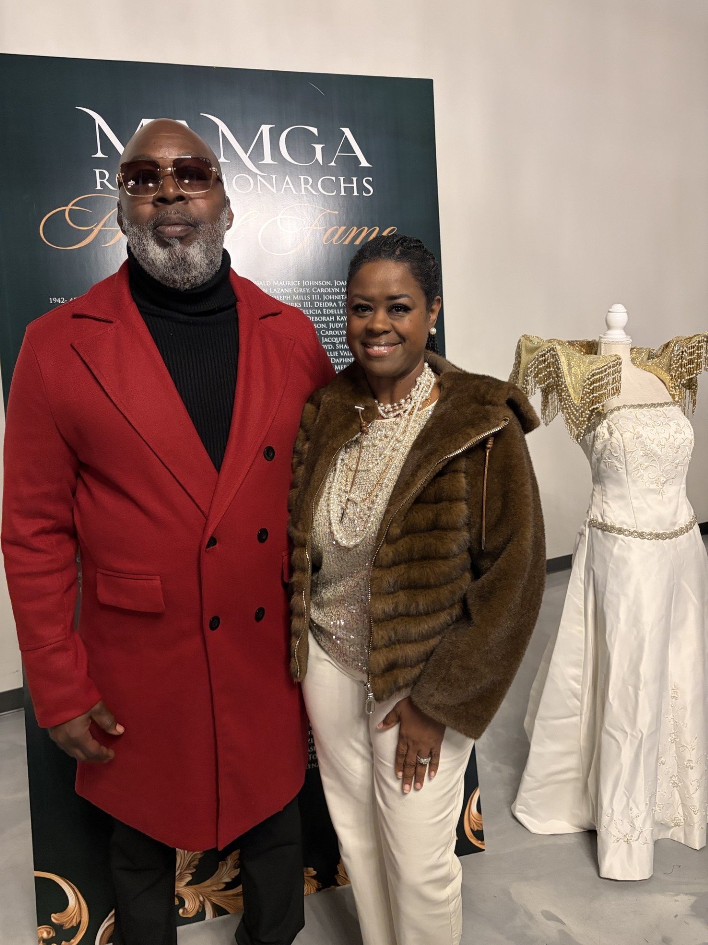 A man and woman standing together, smiling at an event, with a backdrop displaying text about a hall of fame. The man wears a red coat, black shirt, sunglasses, and has a gray beard. The woman wears a brown fur jacket, pearl jewelry, and white pants, with a mannequin dressed in a white gown in the background.