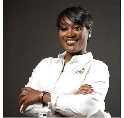 Smiling woman with short black hair, wearing a white collared shirt with a logo, posing with arms crossed against a dark background.