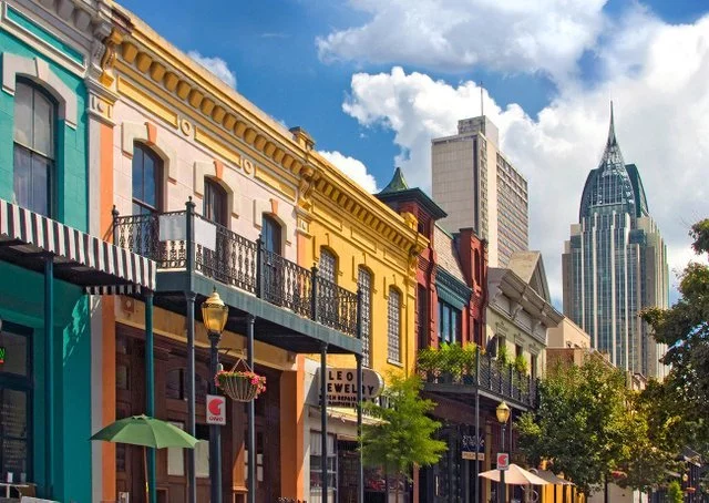 Colorful historic buildings along a street with trees and umbrellas, with modern skyscrapers in the background.
