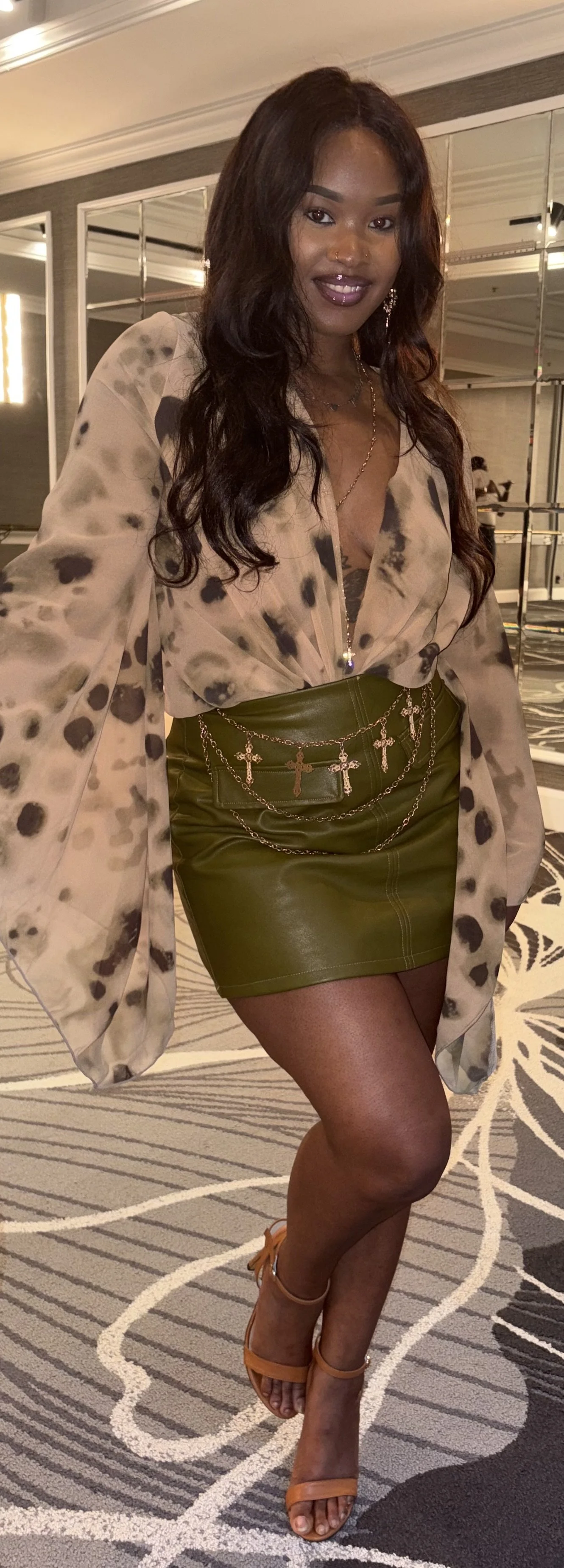 A woman with long, wavy dark hair, wearing a beige and black patterned blouse, a shiny green mini skirt with chain and cross jewelry, and orange high heels, standing on a patterned carpet in a hotel or event space.