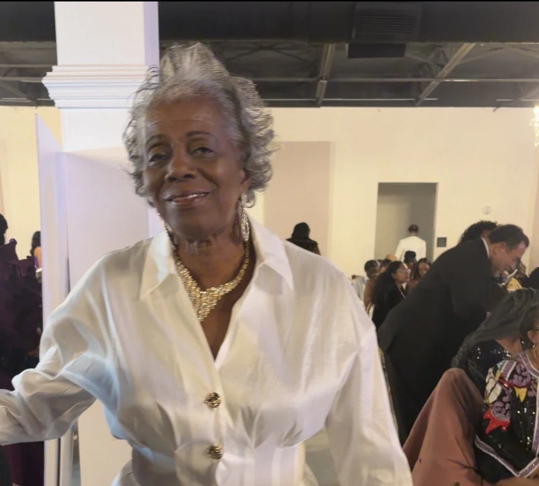 An elderly woman with short gray hair wearing a white blazer and gold jewelry, standing at a social event or gathering.