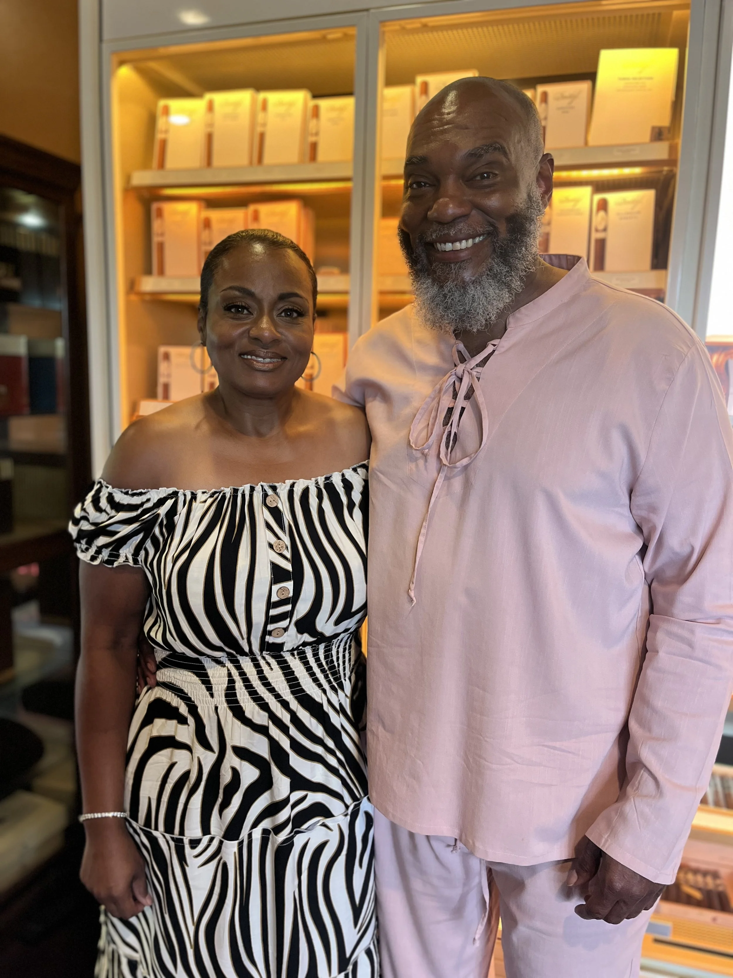 A woman and a man standing close together in a warmly lit room, smiling at the camera. The woman is wearing a black and white striped off-shoulder dress with buttons down the front, and the man is wearing a light pink shirt with a tie at the collar and matching light pink pants. There are bookshelves with yellow lighting behind them.