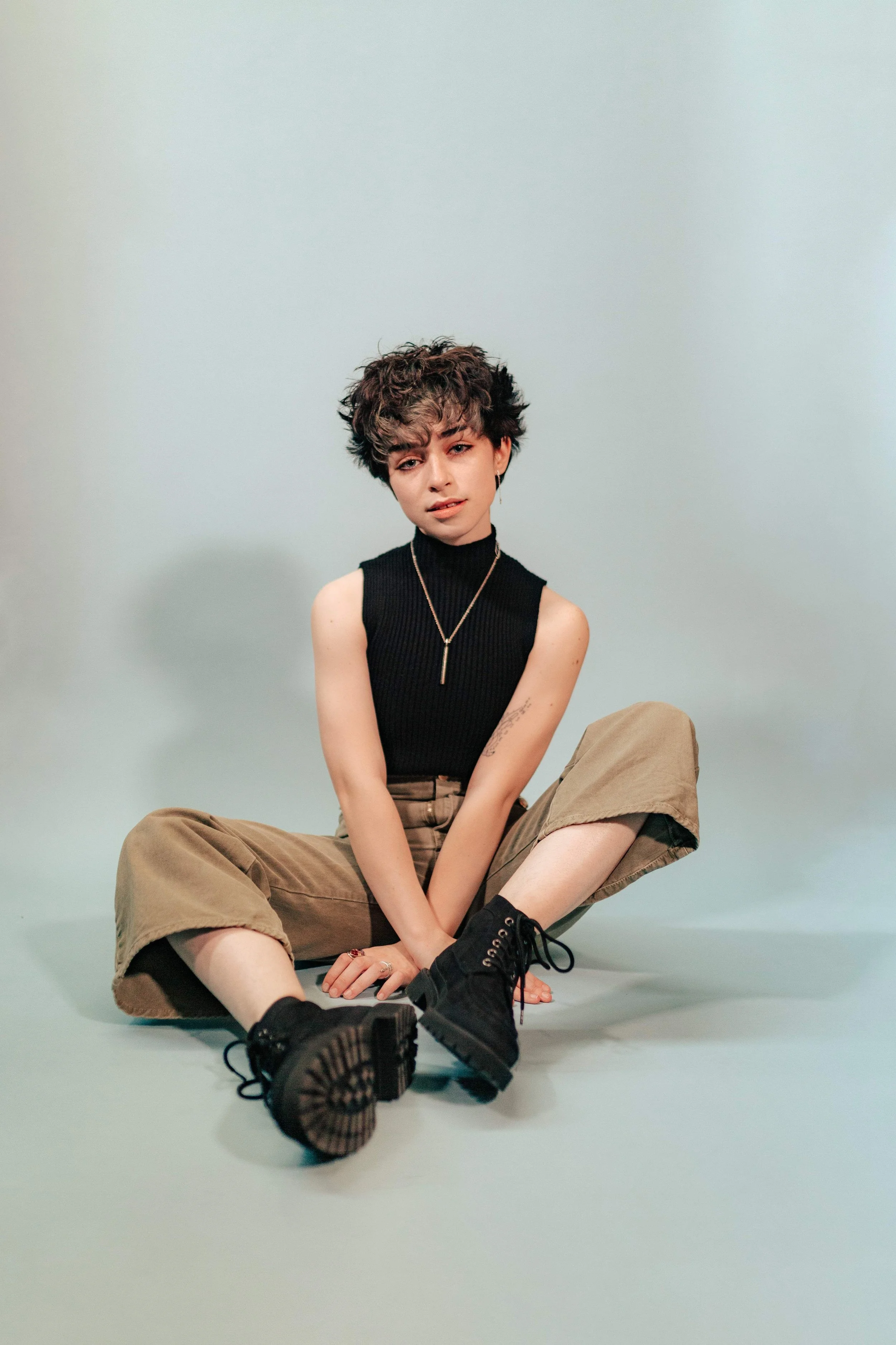 Young woman with short curly hair sitting cross-legged on the floor, wearing a black sleeveless turtleneck top, beige cargo pants, black lace-up boots, and a necklace, against a plain light background.