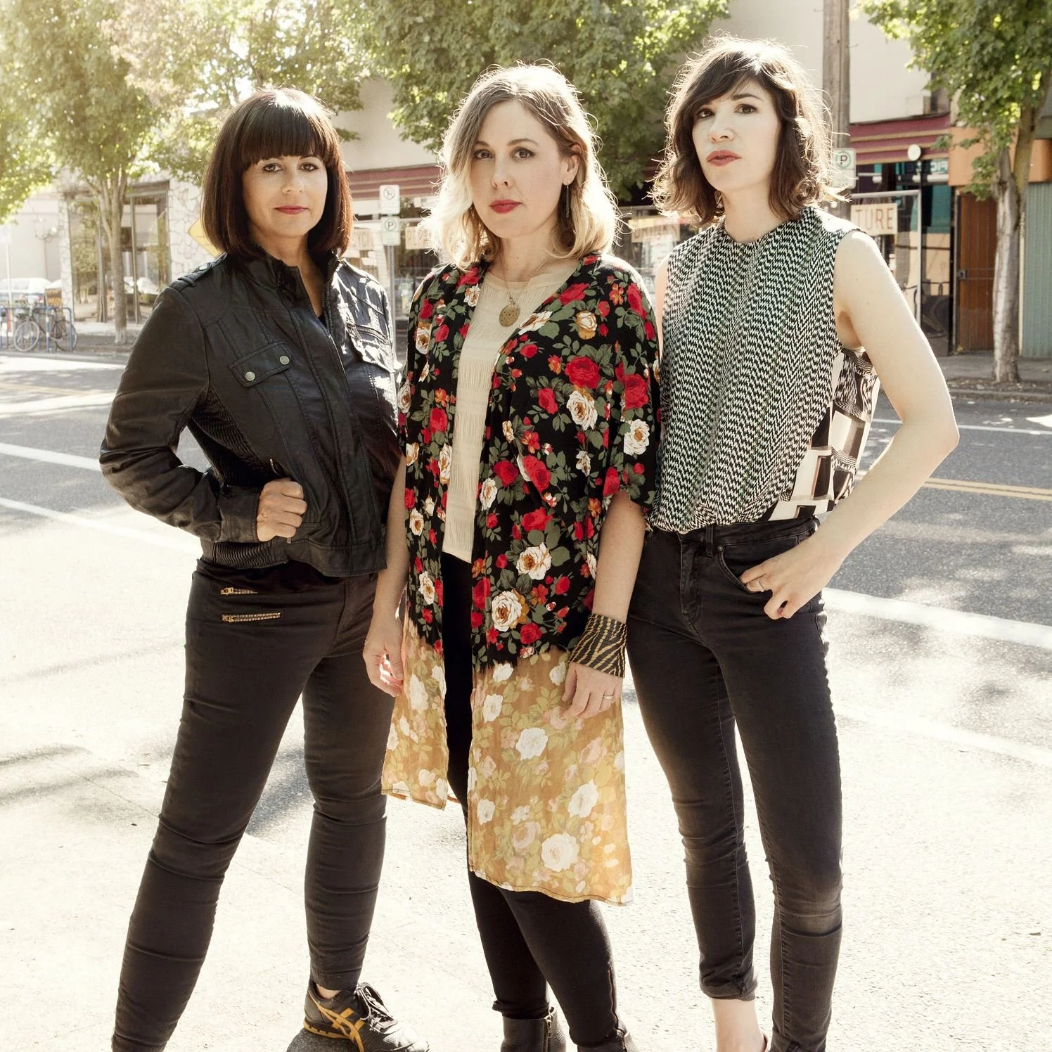 Three women standing on a city street with trees and shops in the background, posing for a photo.