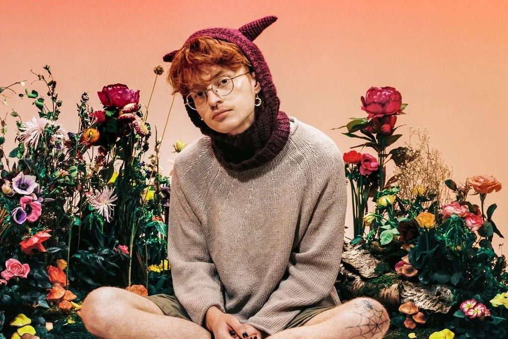 Young person with curly hair, glasses, and tattooed legs sitting among colorful artificial flowers, wearing a beige sweater and a purple knit hat with horns, in front of a pinkish background.