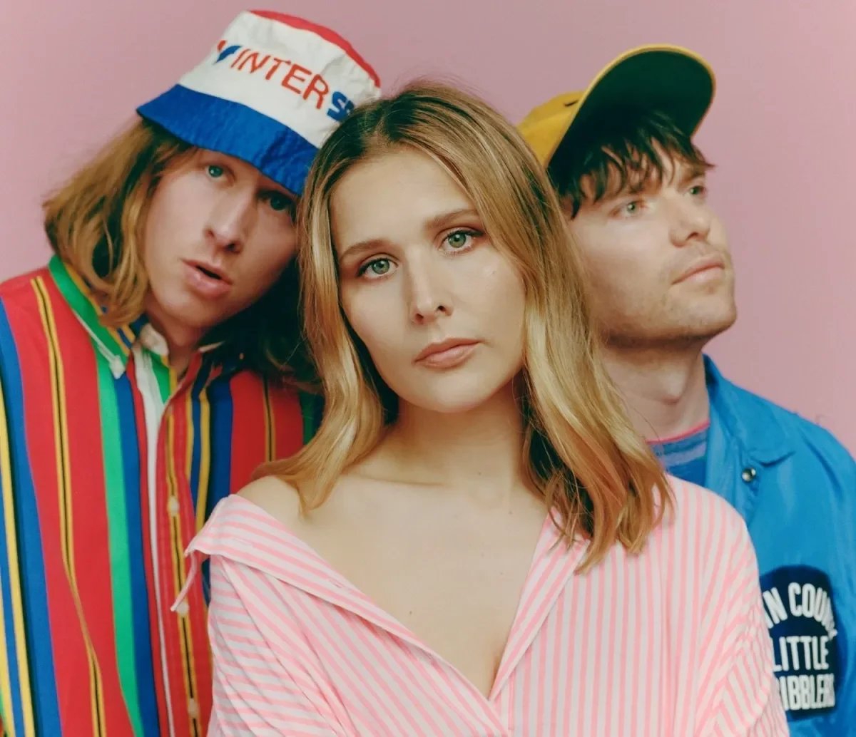 Three young adults, two men and one woman, posing against a pink background. The woman has blonde hair and is wearing a pink and white striped shirt. The man on the left has reddish hair, a striped jacket, and a hat that says 'INTER'. The man on the right has dark hair, wears a yellow cap and a blue jacket.