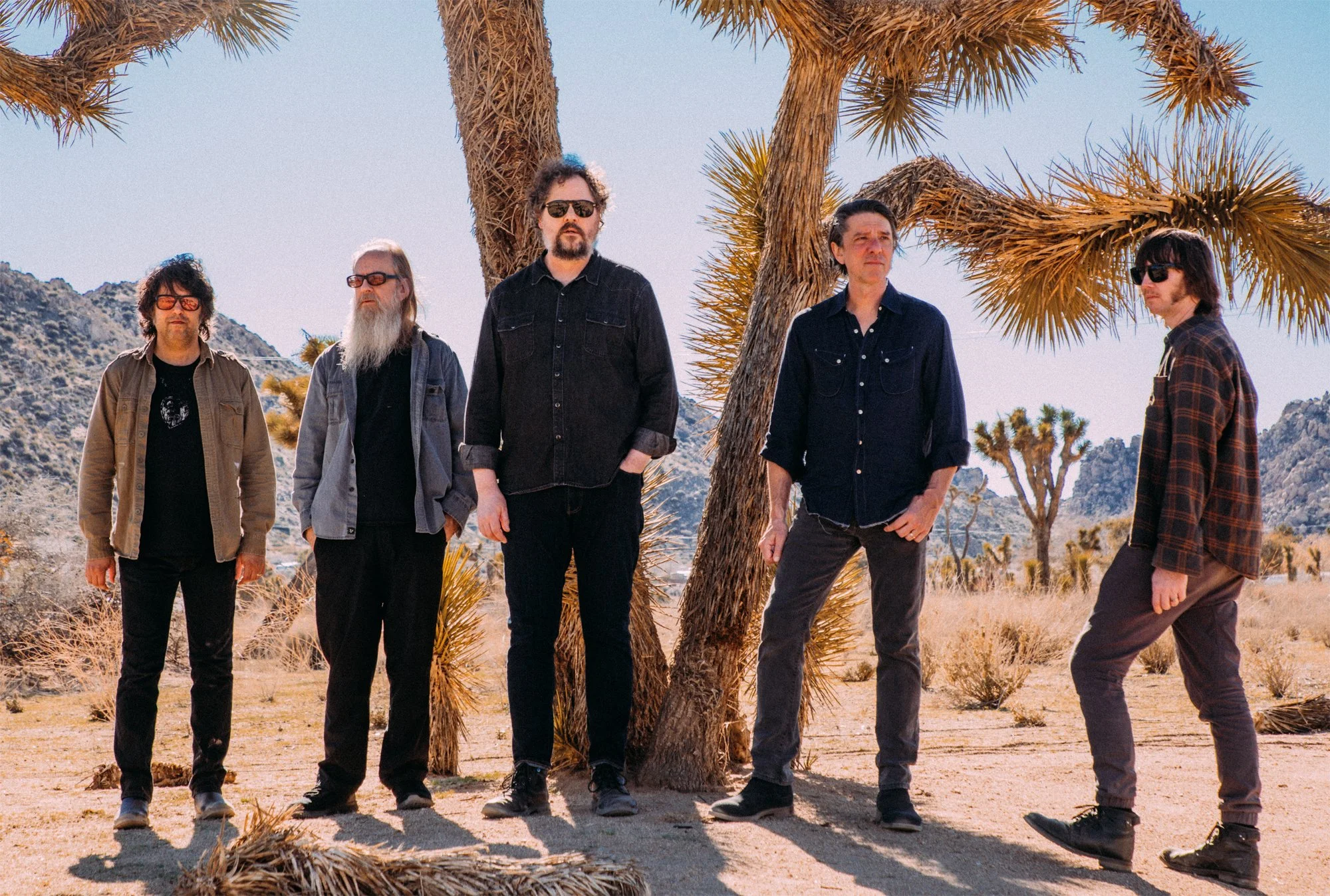 Five men standing in a desert landscape with Joshua trees and mountains in the background. They are dressed casually and looking in different directions.