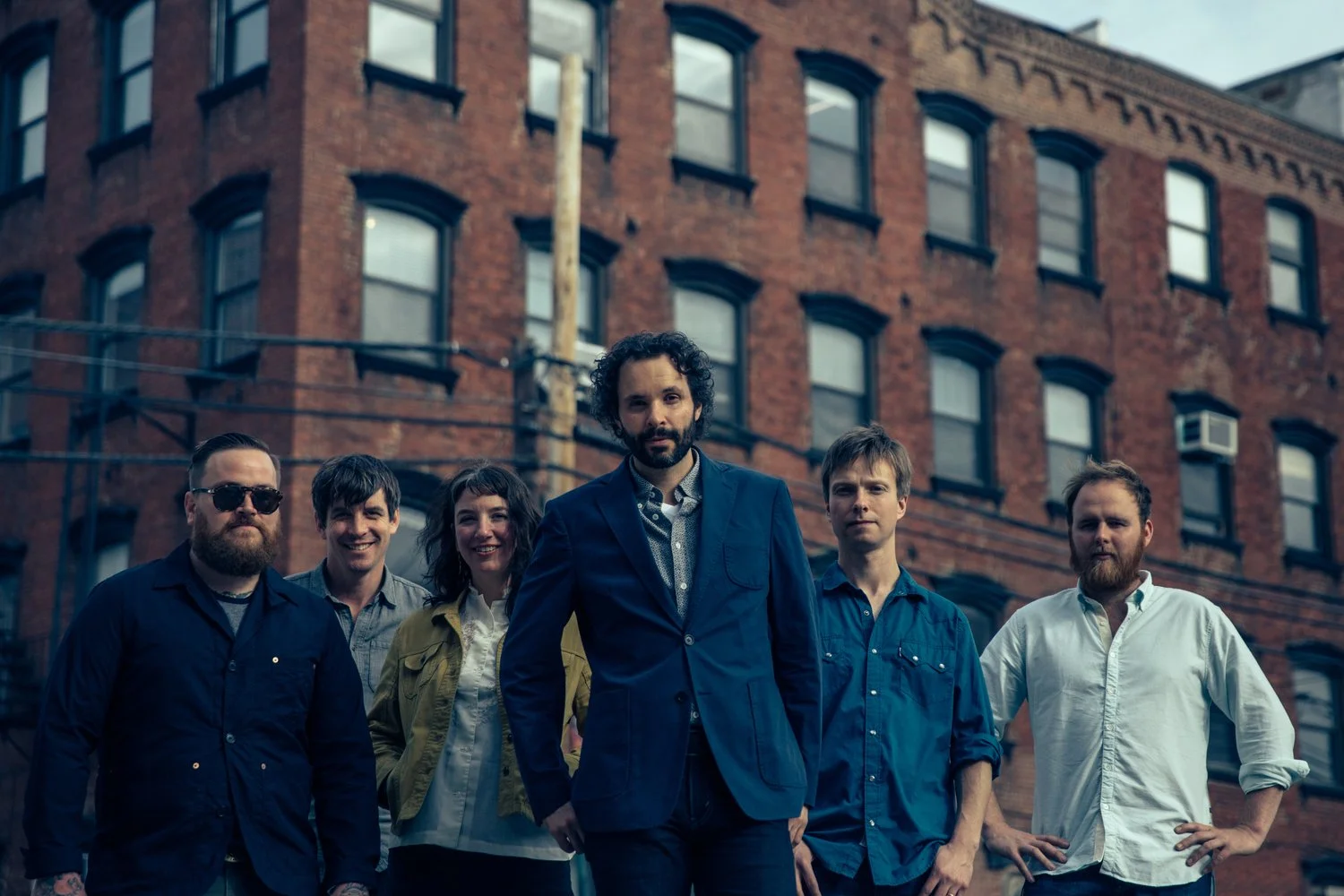 Group of six people standing outdoors in front of a brick building, some with their hands on their hips, posing for a photo.