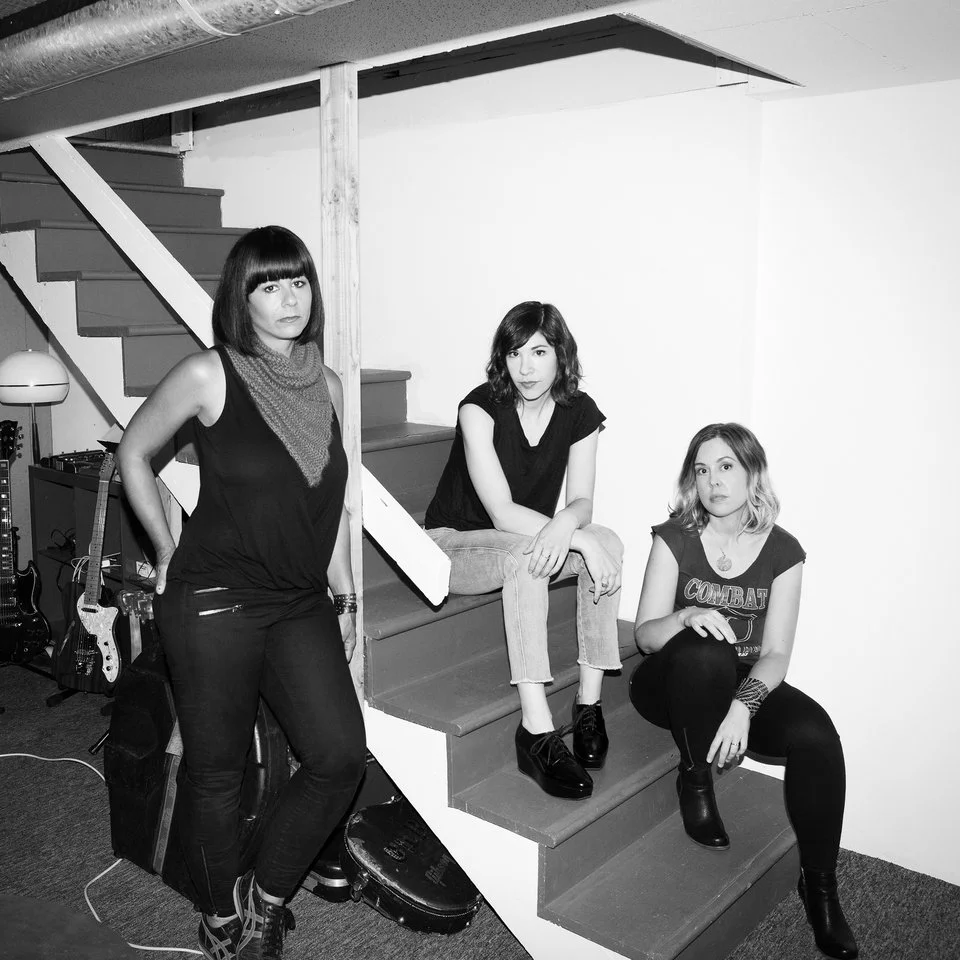 Three women with dark hair and casual clothing sitting and standing on a staircase in a room with musical instruments and equipment.
