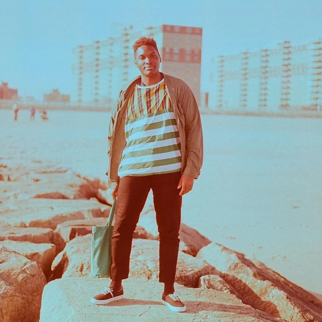 A young man standing on rocks near a body of water with city buildings in the background, wearing a colorful striped shirt, dark pants, a brown jacket, and holding a tote bag.