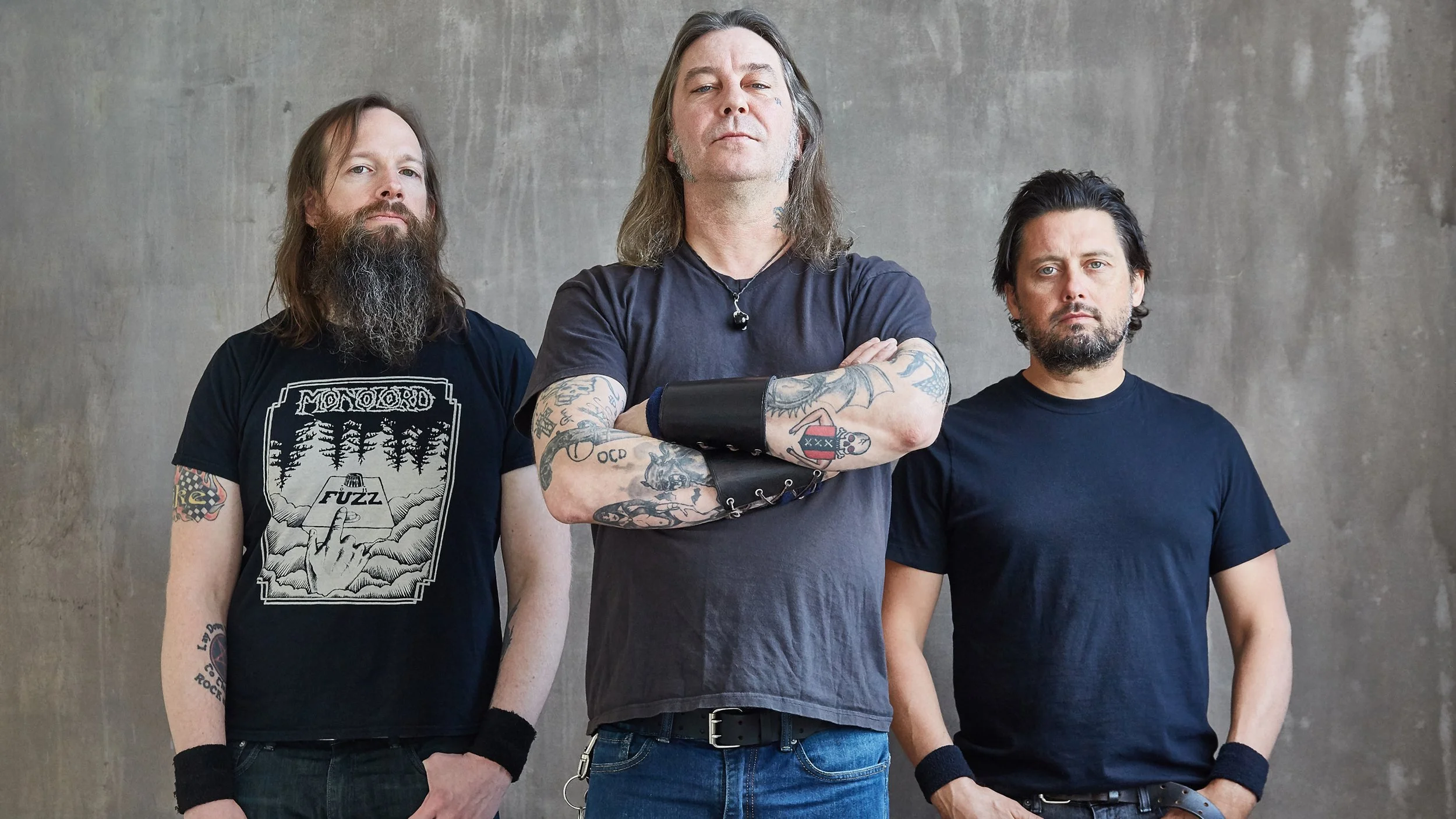 Three men standing side by side against a plain wall, each with arms crossed or at their sides, wearing dark t-shirts, with the man in the center having long hair and tattoos on his arms.