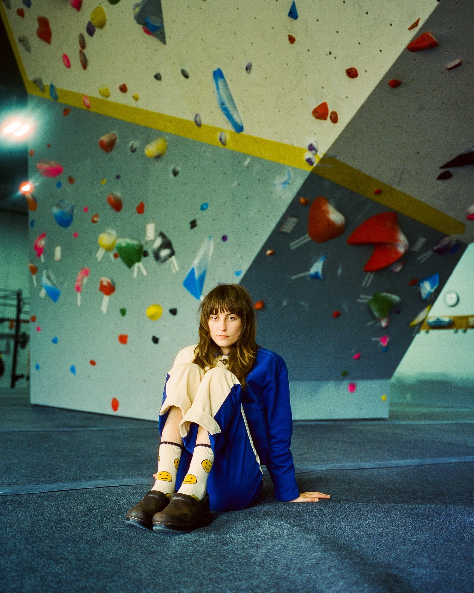 A young woman with brown hair sitting on the floor in an indoor climbing gym, with a large, colorful climbing wall in the background.