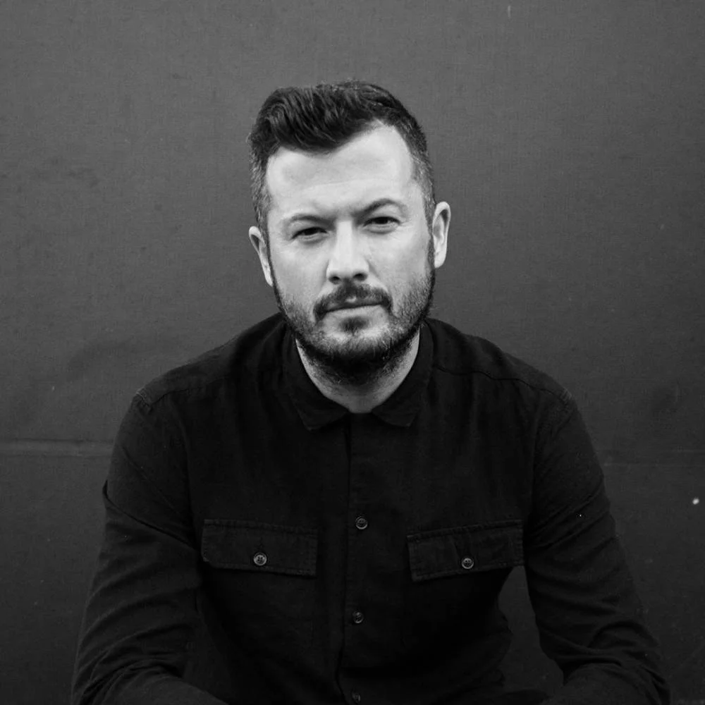 A black-and-white photo of a man with short hair and a beard, wearing a dark button-up shirt, sitting against a plain dark background.