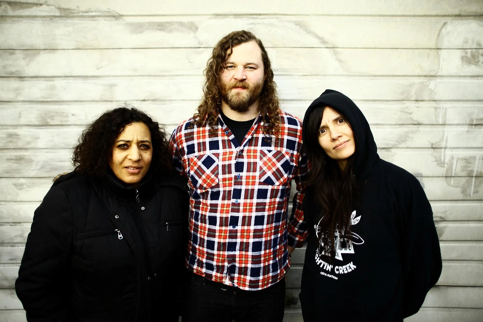 Three people standing in front of a gray wooden wall, two women on either side of a man with long curly hair and a beard, wearing casual clothing.