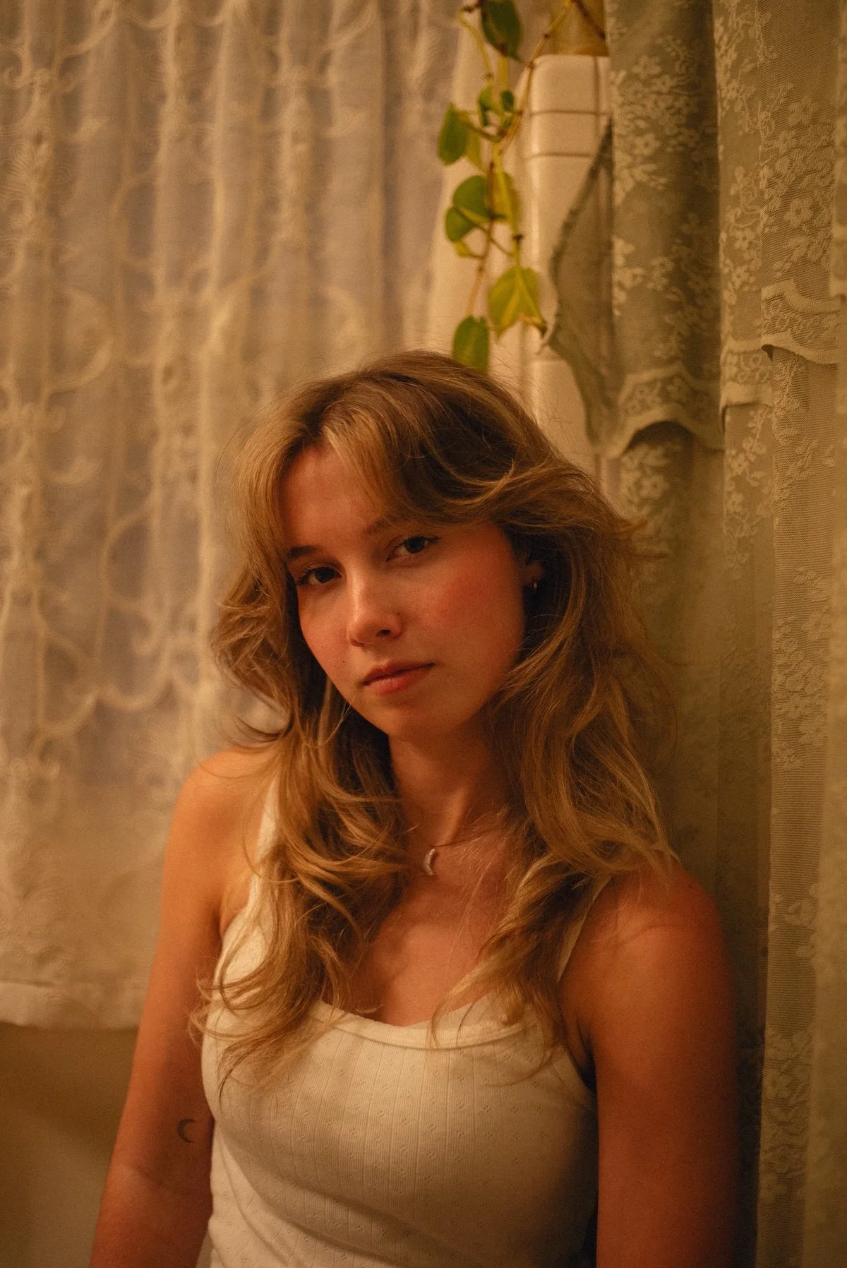 A young woman with wavy blonde hair wearing a white tank top, sitting indoors in front of lace curtains and a leafy plant. Warm lighting.
