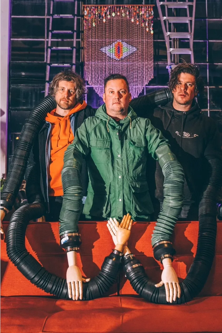 Three men standing behind a red table with robotic arms attached to their lower arms, in front of a backdrop with stage lighting and a digital art piece hanging.