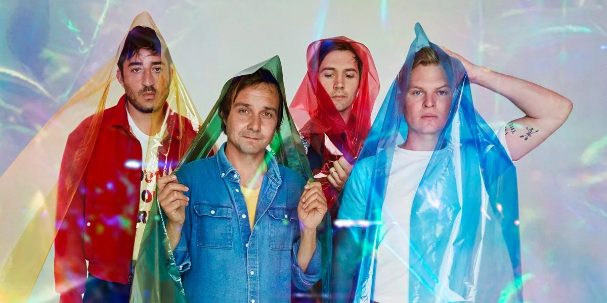 Four young men standing together, each wearing a colorful translucent raincoat, with a light, rainbow-like background.