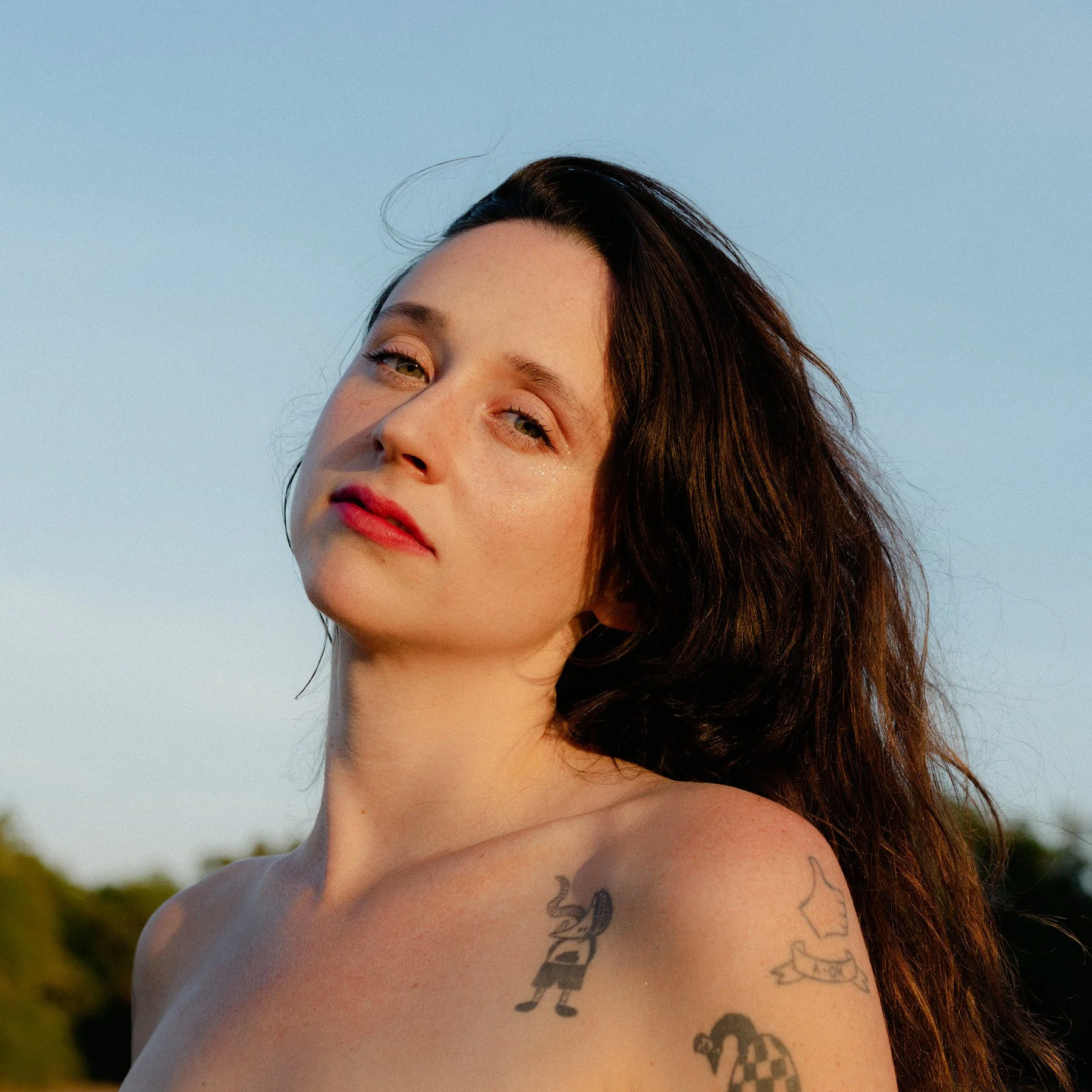 A woman with long dark hair and tattoos on her shoulder outdoors during sunset, looking at the camera with a neutral expression.
