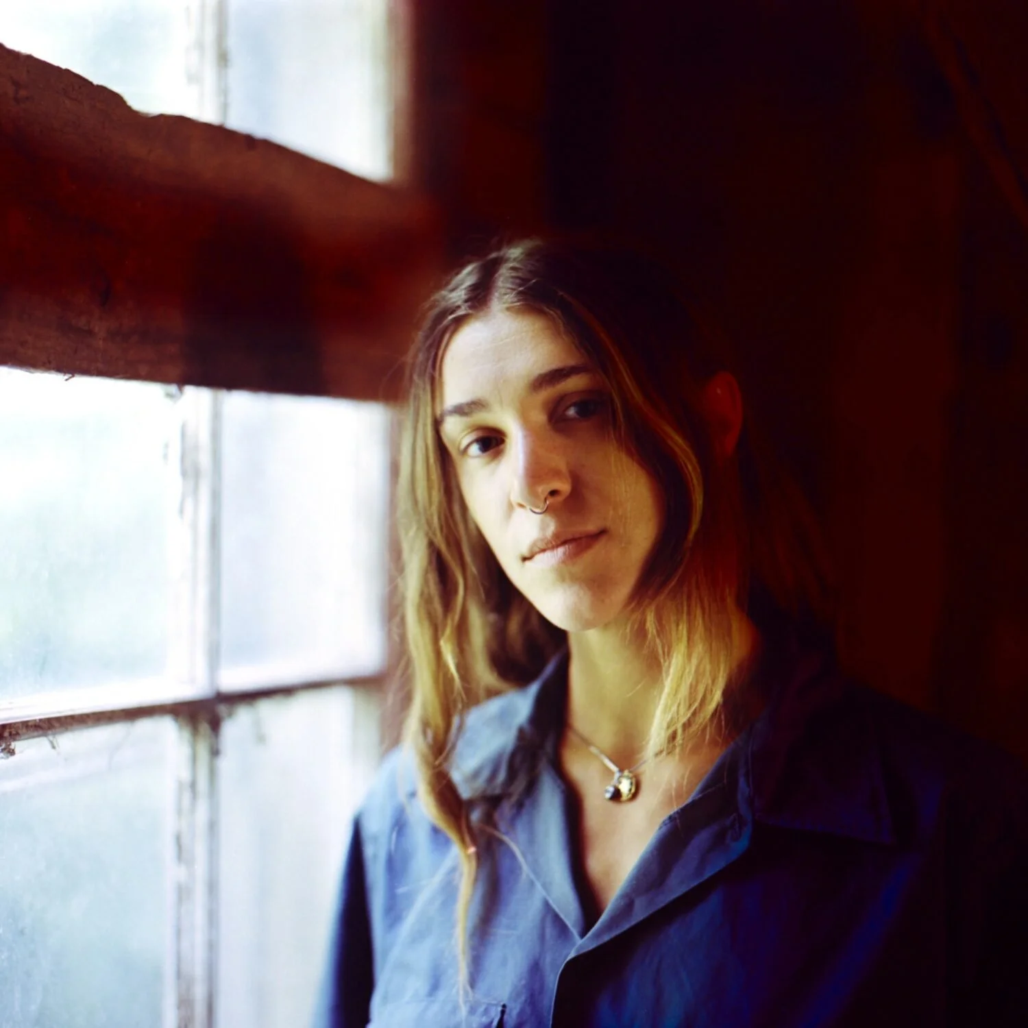 A young woman with shoulder-length light brown hair and a septum piercing looks at the camera by a frosted window with wooden framing, wearing a denim shirt and a necklace, inside a dimly lit room.