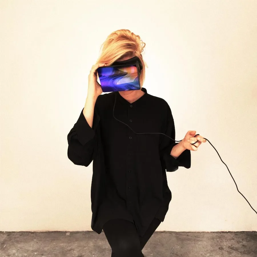 Person with short blonde hair and black clothing wearing a virtual reality headset and holding a controller.