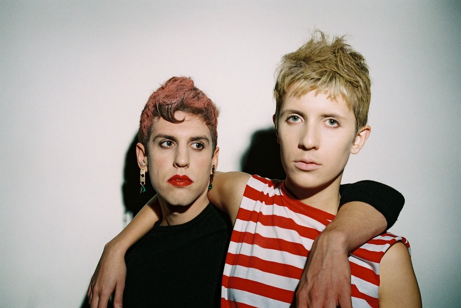 A woman with short reddish hair and red lipstick is standing next to a young man with short blonde hair wearing a sleeveless red and white striped shirt. The woman has her arm around the young man's shoulder, and they are both looking directly at the camera with serious expressions.