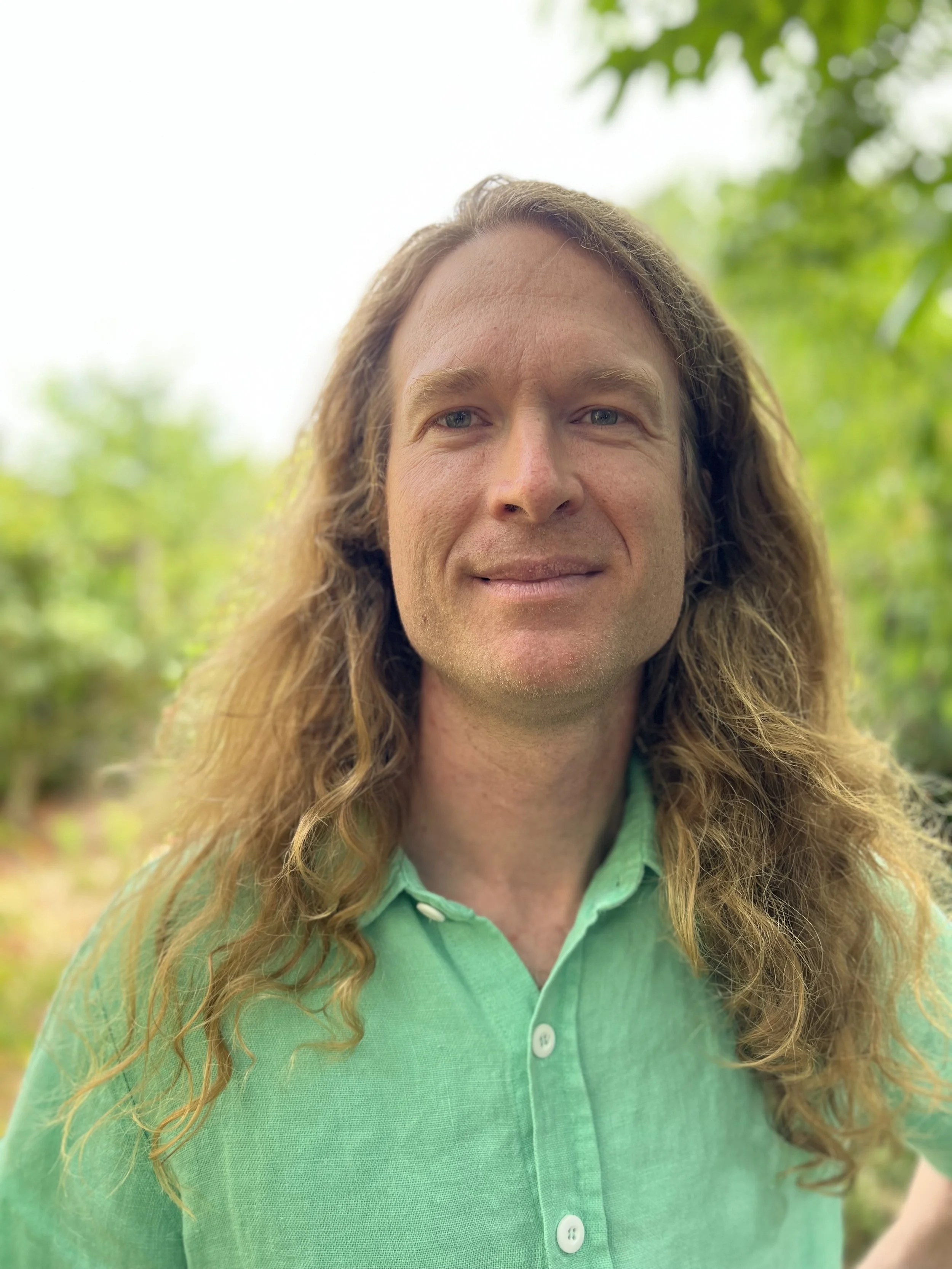 A man with long, wavy hair wearing a green button-up shirt outdoors with blurred green foliage in the background.