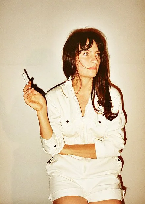 A woman with long dark hair wearing a white shirt and shorts, holding a cigarette in one hand and looking to the side.