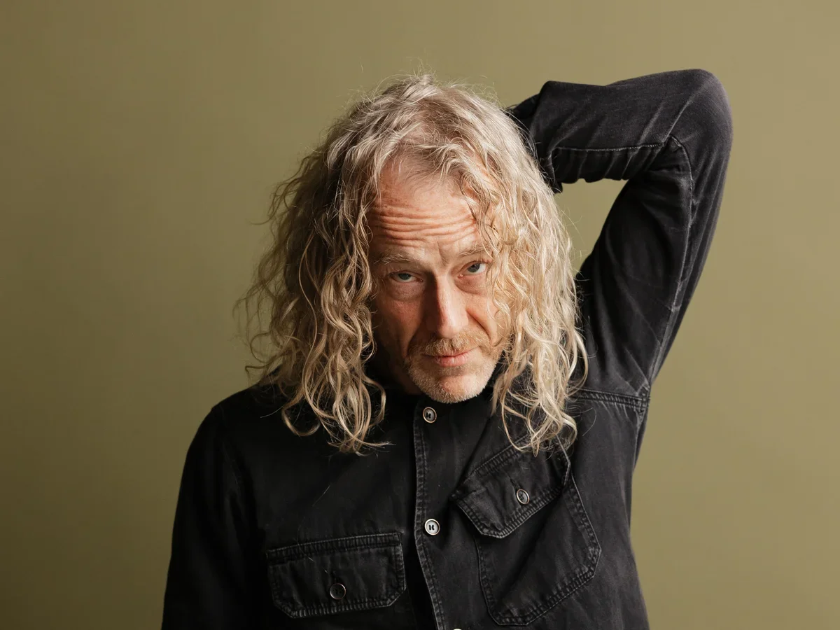 A man with long, wavy blond hair and a light beard, wearing a black denim shirt, looking at the camera with his right arm raised behind his head, against a plain olive green background.