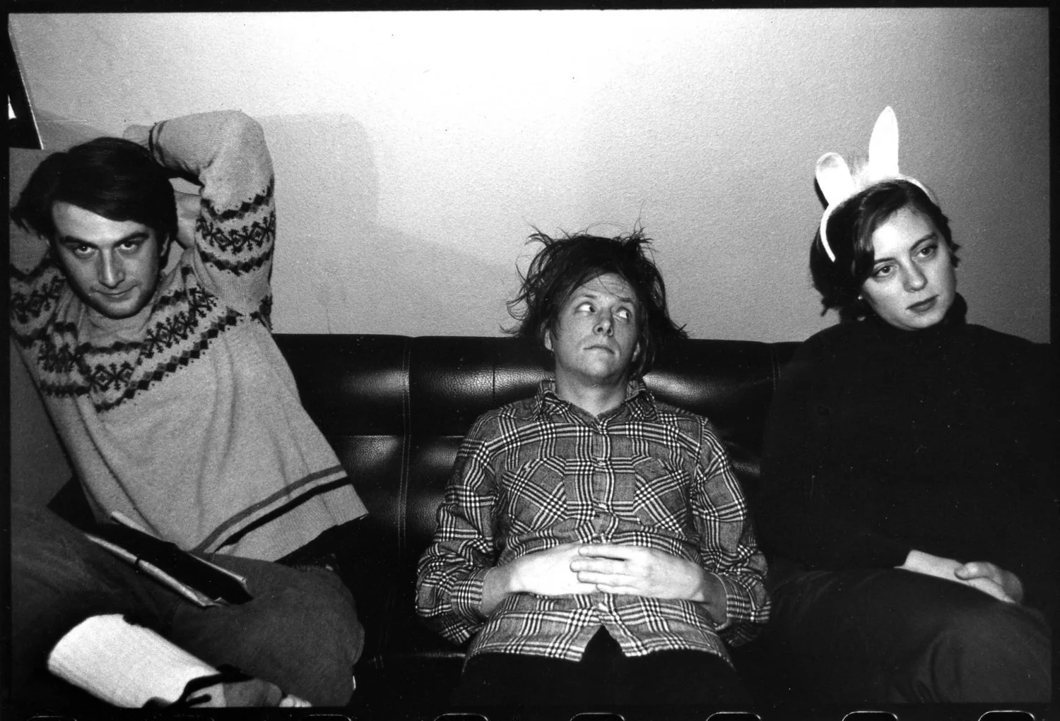 Three people sitting on a couch, with one person in the middle looking confused or tired, wearing a plaid shirt, the person on the left in a patterned sweater with their hands behind their head, and the person on the right with a headband with bunny ears, wearing a dark top.