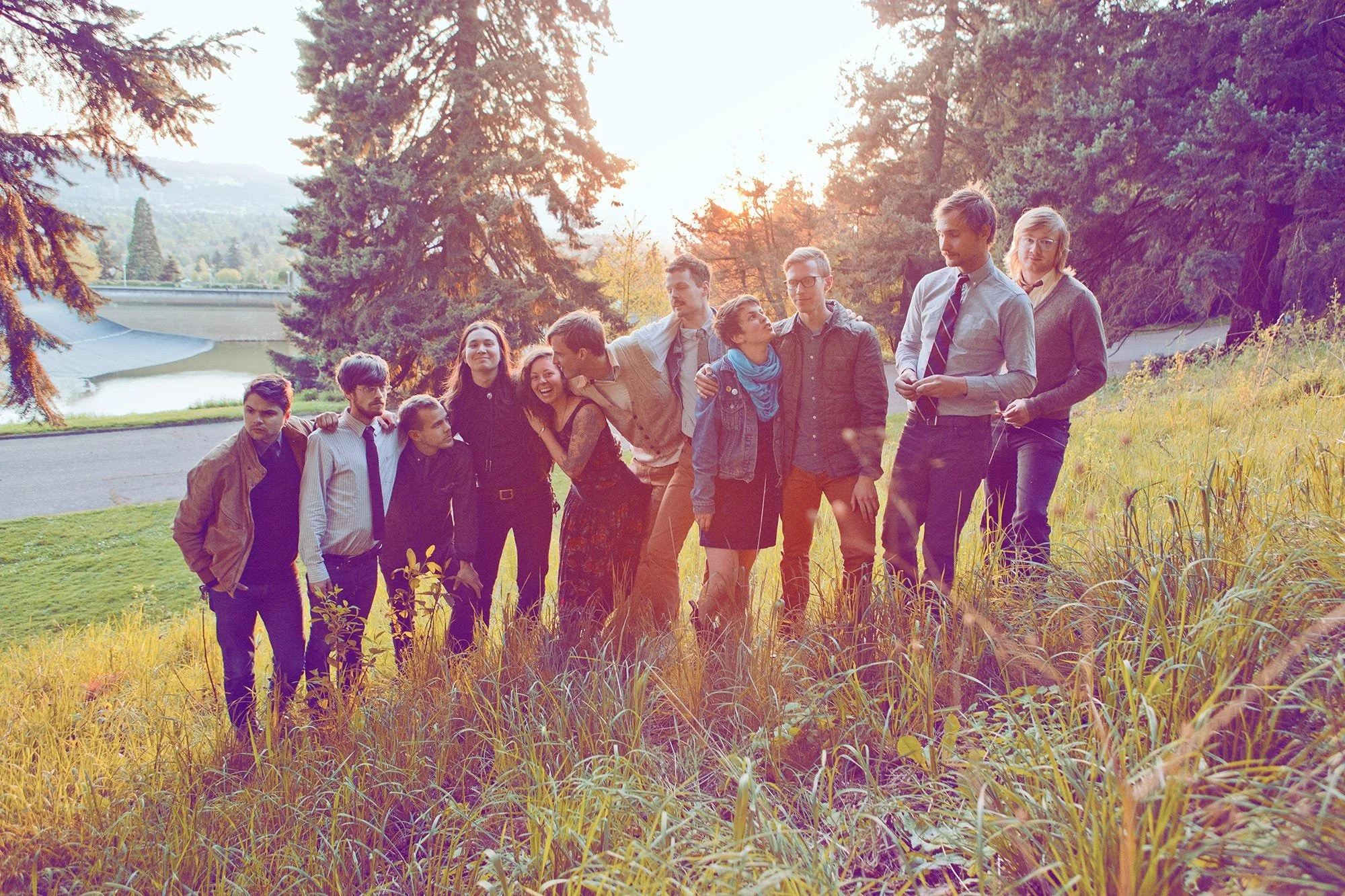 Group of people standing together outdoors during sunset in a park with trees and a pond.