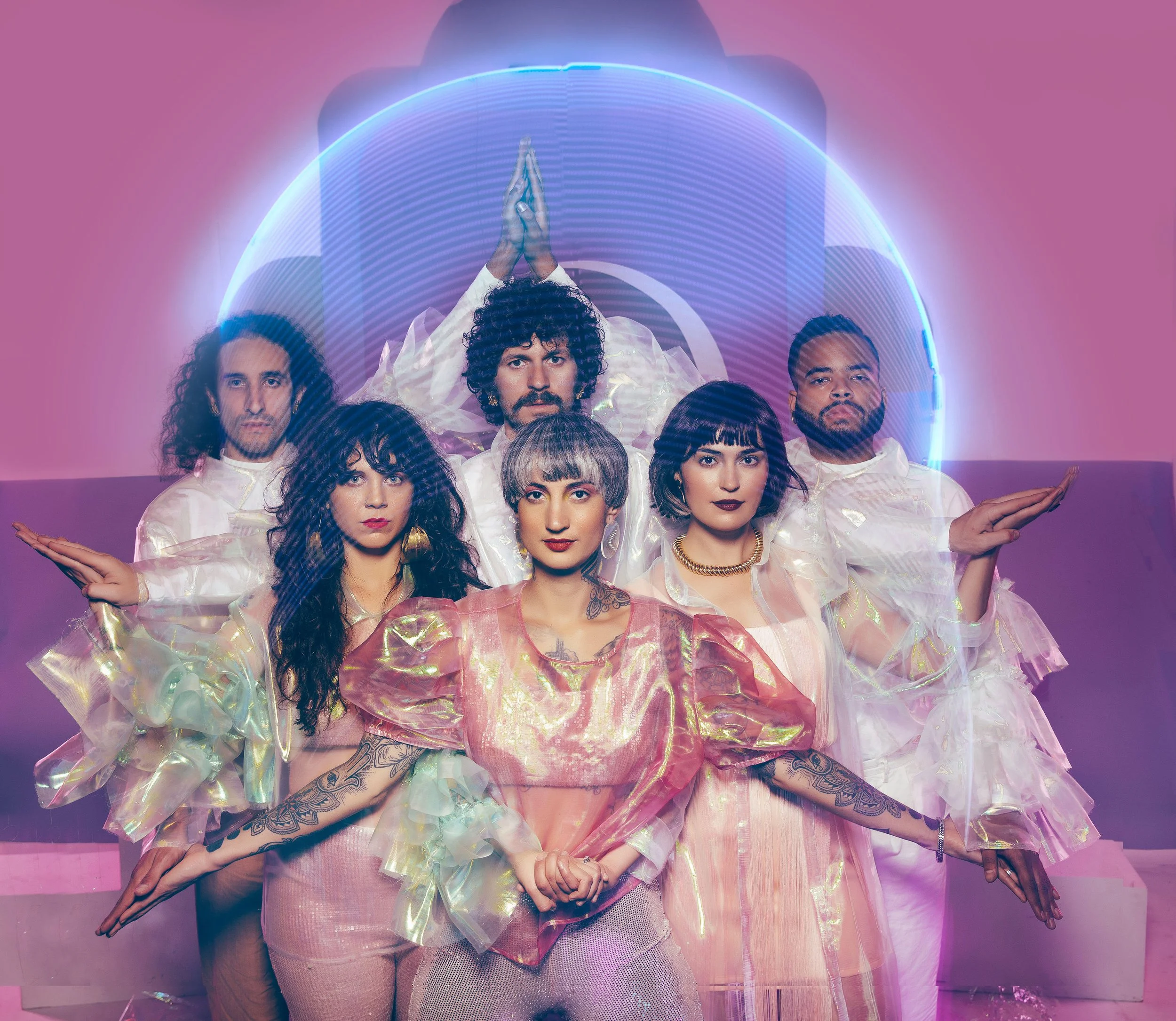 A group of six people dressed in iridescent, pastel-colored outfits, standing in front of a neon blue halo with a pink background, with three people in the front row and three behind, all striking confident poses with calm expressions.