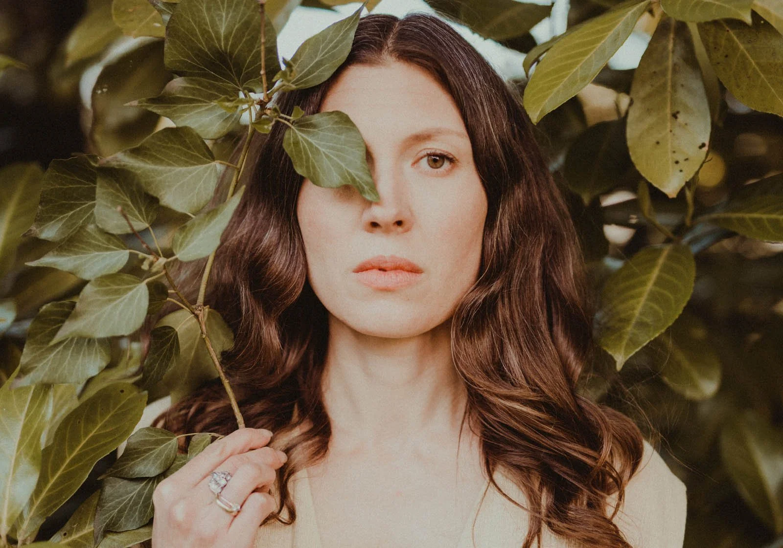A woman with long, wavy brown hair standing among large green leaves, with one leaf partially covering her eye, looking directly at the camera with a neutral expression.
