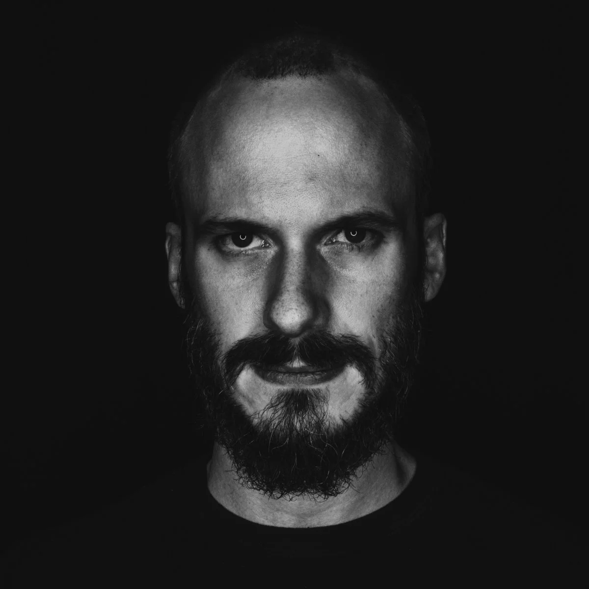 Black and white portrait of a man with a beard and mustache, looking directly at the camera, against a dark background.